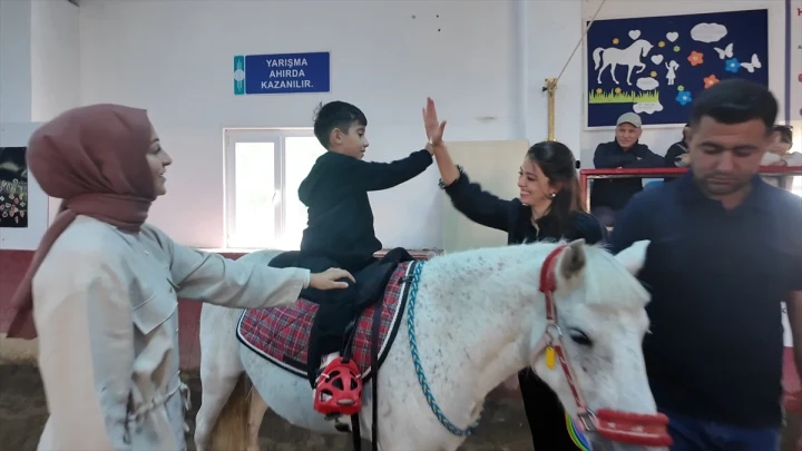 Hippoterapi merkezinde at üzerinde oturan genç bir katılımcı, yanında duran bir kadınla el çakışıyor.