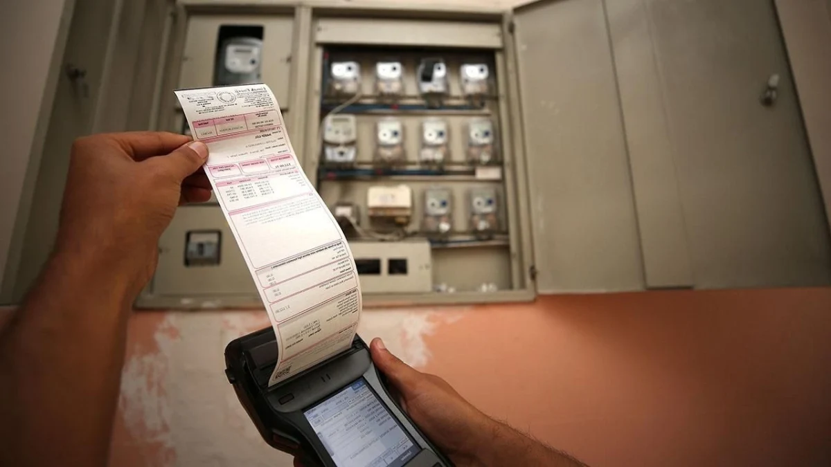 Elektrik sayaçlarının önünde el terminalinden çıkan tüketim fişini tutan kişi
