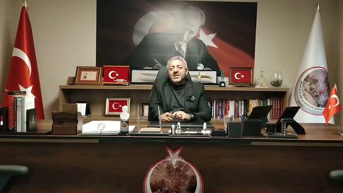 Masa başında oturan bir kişi; arkasında Türk bayrakları ve Atatürk portresi bulunuyor.