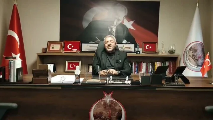 Masa başında oturan bir kişi; arkasında Türk bayrakları ve Atatürk portresi bulunuyor.