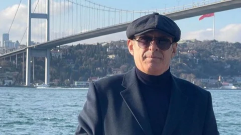 Koyu renk şapka ve gözlük takan bir erkek, deniz ve asma köprü önünde oturuyor.