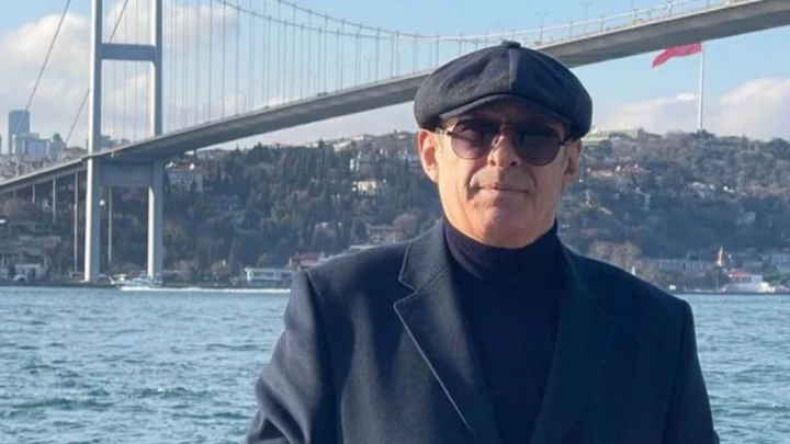 Koyu renk şapka ve gözlük takan bir erkek, deniz ve asma köprü önünde oturuyor.