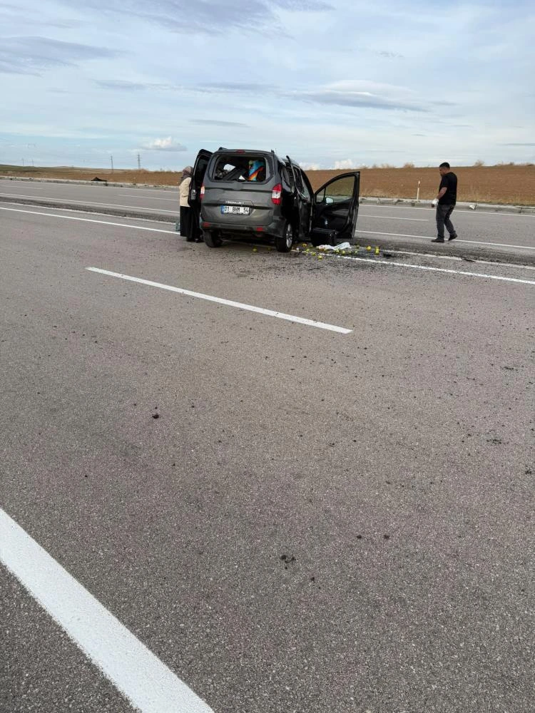 Yol kenarında durmuş, hasar görmüş koyu renkli otomobil ve çevresindeki kişiler