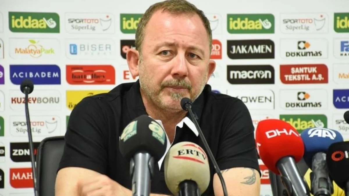 Sergen Yalçın, sponsor panosu önünde mikrofonların karşısında oturuyor.