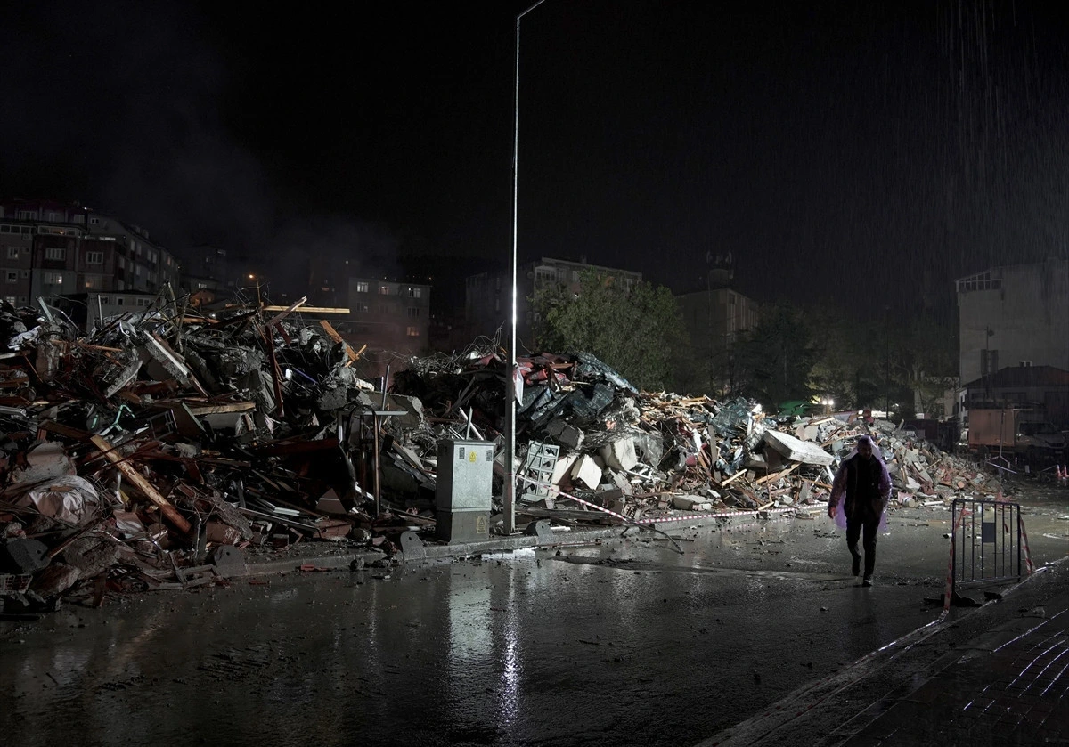 Gece yağmur altında yıkıntıların bulunduğu alanda yürüyen bir kişi görülüyor.